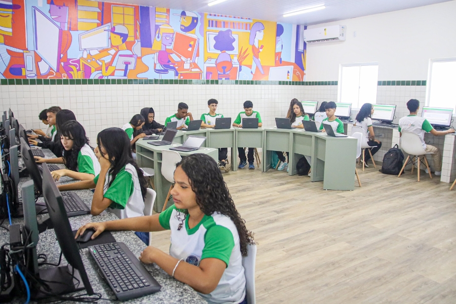 Sala de informática com piso de madeira clara, paredes de azulejo branco e um painel colorido com ilustrações de tecnologia. Cerca de 18 estudantes, em uniforme branco e verde, estão concentrados, alguns em computadores de mesa e outros em notebooks, dispostos em mesas cinza claro. A sala é bem iluminada, com janelas e ar-condicionado
