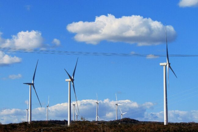 A imagem mostra diversos aerogeradores (turbinas eólicas) espalhados por uma paisagem de vegetação seca, sob um céu azul com nuvens brancas