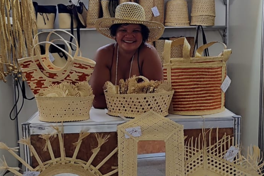 Mulher sorridente com chapéu de palha posa atrás de uma mesa repleta de artesanato feito com palha, como bolsas, enfeites e cestos. Ao fundo, prateleiras exibem mais peças trançadas cuidadosamente