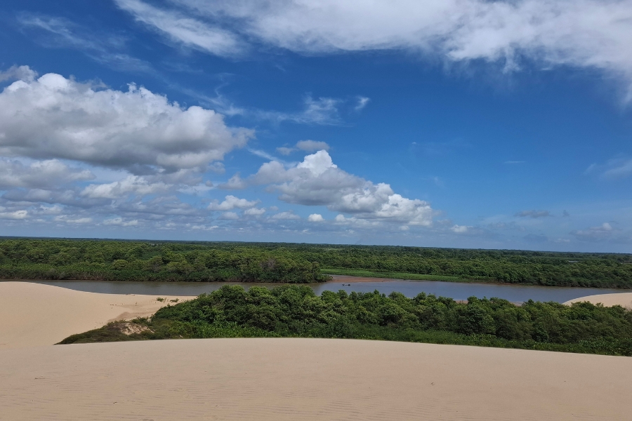 A imagem mostra uma paisagem natural com dunas de areia clara em primeiro plano, um rio sinuoso ao centro e uma densa faixa de mata verde ao fundo, sob um céu azul com algumas nuvens brancas