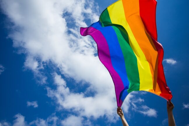 Bandeira do arco-íris, símbolo do orgulho LGBTQIAPN+, é erguida por duas mãos contra um céu azul com nuvens brancas. O tecido colorido balança ao vento, destacando-se em contraste com o fundo ensolarado