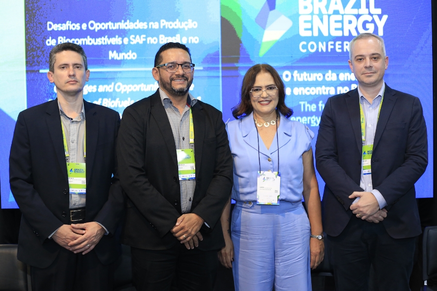 Quatro pessoas — três homens e uma mulher — estão lado a lado, posando para foto em um evento. Estão vestidos com roupas sociais e usam crachás. Ao fundo, um painel azul exibe o texto “Brazil Energy Conference” e informações sobre produção de biocombustíveis