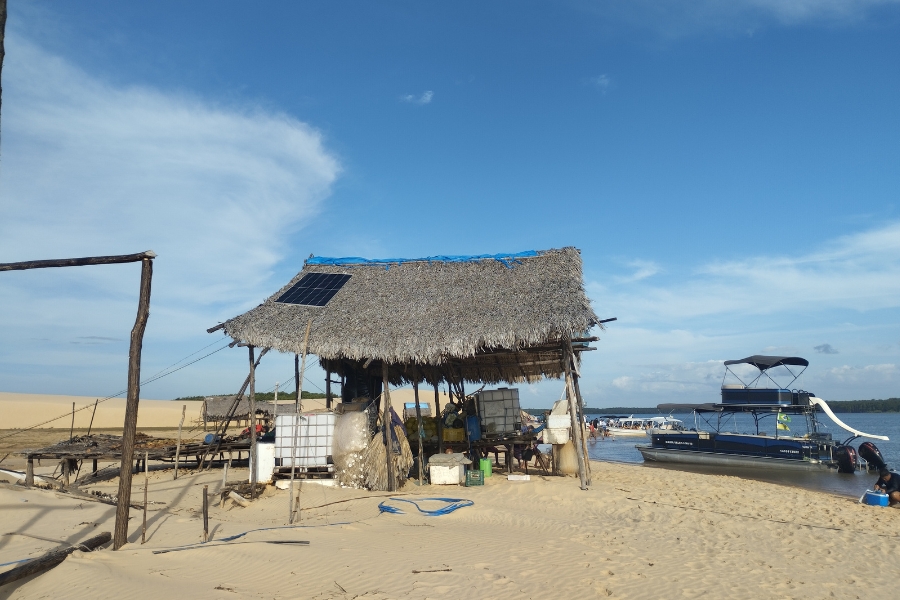 A foto mostra um rancho rústico de pescadores, com cobertura de palha e placas solares no telhado, em uma praia de areia clara. Ao lado, há um barco atracado na beira do rio sob um céu azul