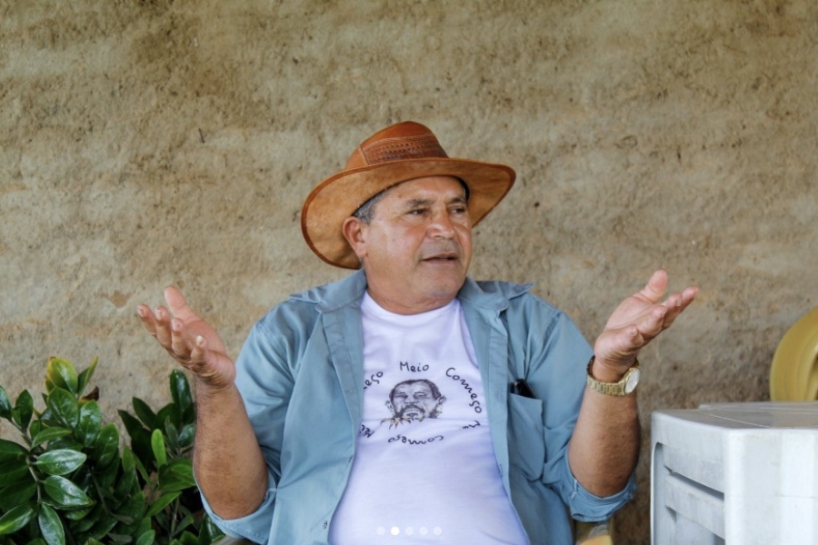 Homem de pele clara e idade madura, com chapéu de couro marrom, camiseta branca com estampa e camisa azul aberta por cima. Ele está sentado, gesticulando com as mãos abertas, em um ambiente rústico de parede de barro. Há plantas e mesas plásticas empilhadas ao lado