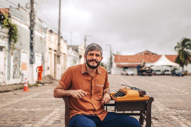 A imagem mostra um homem sentado em uma cadeira no meio de uma rua de paralelepípedos, em uma área urbana de aparência histórica. Ele tem cabelo grisalho curto, barba e bigode, e usa óculos de armação laranja. Está vestido com uma camisa marrom de mangas curtas e calça azul. À sua frente, sobre uma pequena mesa de madeira, há uma máquina de escrever antiga, de cor laranja vibrante. Ao fundo, é possível ver casas antigas com fachadas desgastadas, um céu nublado e uma tenda branca ao lado de carros estacionados. A expressão do homem é tranquila