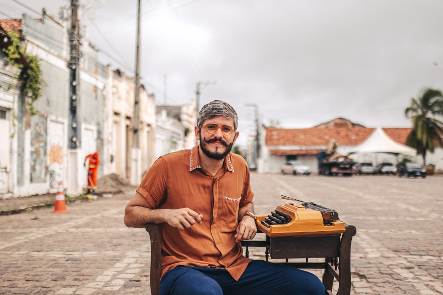 A imagem mostra um homem sentado em uma cadeira no meio de uma rua de paralelepípedos, em uma área urbana de aparência histórica. Ele tem cabelo grisalho curto, barba e bigode, e usa óculos de armação laranja. Está vestido com uma camisa marrom de mangas curtas e calça azul. À sua frente, sobre uma pequena mesa de madeira, há uma máquina de escrever antiga, de cor laranja vibrante. Ao fundo, é possível ver casas antigas com fachadas desgastadas, um céu nublado e uma tenda branca ao lado de carros estacionados. A expressão do homem é tranquila