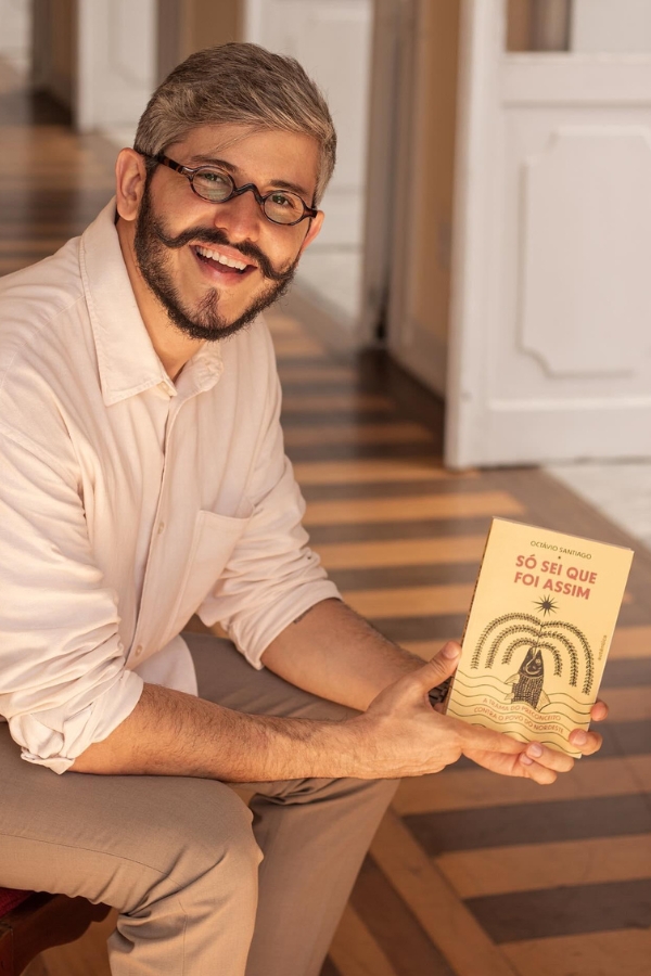 A imagem mostra um homem sorridente sentado em um ambiente interno iluminado pela luz do sol. Ele tem cabelo grisalho curto, barba e bigode modelado com as pontas enroladas para cima, e usa óculos de armação redonda escura. Está vestido com uma camisa social clara e calça bege. Ele segura um livro com as duas mãos, mostrando a capa para a câmera. O título do livro é “Só sei que foi assim”, de autoria de Octavio Santiago. A capa tem ilustrações simples em preto e tons terrosos, com uma figura de cangaceiro e elementos da cultura nordestina. O chão tem um padrão geométrico de madeira em tons escuros e claros, e ao fundo há portas brancas abertas