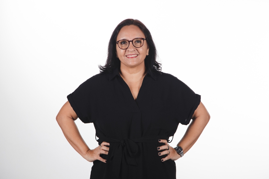 Foto de uma mulher de pele clara e cabelos escuros, lisos e na altura dos ombros. Ela usa óculos de armação escura e está sorrindo, olhando diretamente para a câmera. Veste um vestido preto de manga curta com um cinto da mesma cor, amarrado na cintura. Suas mãos estão apoiadas na cintura. Ao fundo, o cenário é neutro, com fundo branco liso