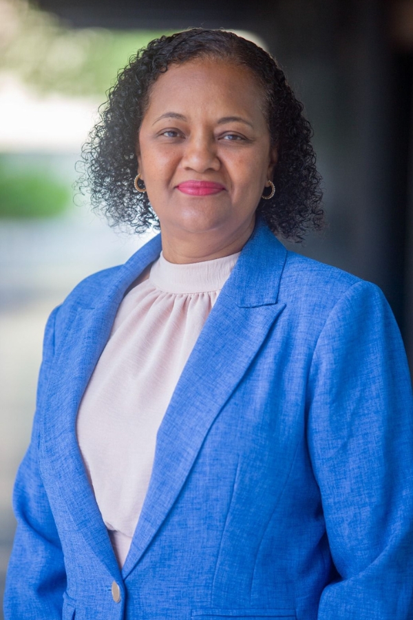 A foto mostra uma mulher de pele morena clara e cabelos curtos, crespos e pretos, que sorri levemente para a câmera. Ela veste um blazer azul vibrante sobre uma blusa clara de gola arredondada. Ao fundo, a imagem está desfocada, com tons neutros e um pouco de verde