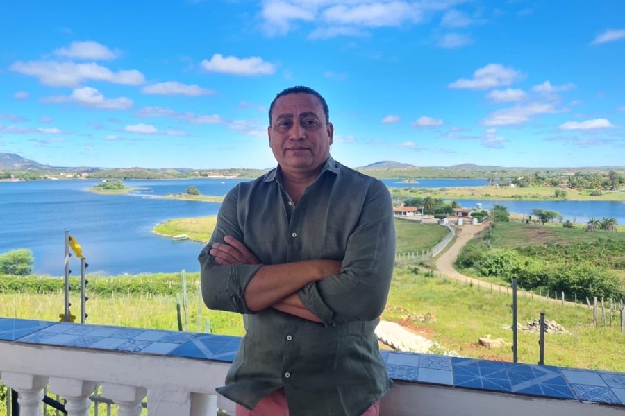A foto mostra um homem de pele morena e cabelos curtos, vestindo uma camisa verde-escura de mangas compridas e calça clara, em pé com os braços cruzados. Ele está em uma varanda de azulejos azuis e brancos, tendo ao fundo uma paisagem ampla com um lago de águas azuis, pequenas ilhas verdes, casas espalhadas e uma estrada de terra que serpenteia pelo campo. O céu está claro e azul, pontuado por algumas nuvens brancas