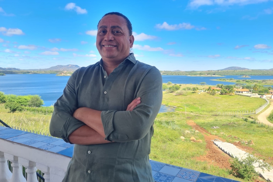 A imagem mostra um homem de pele morena e cabelos escuros e curtos, sorrindo e olhando para a câmera. Ele veste uma camisa de manga longa verde-oliva, com os braços cruzados à frente do corpo. Ao fundo, há uma paisagem ampla e ensolarada: um grande lago de águas azuis, áreas de vegetação verde e algumas casas espalhadas. Montanhas baixas aparecem no horizonte, sob um céu azul claro com poucas nuvens brancas