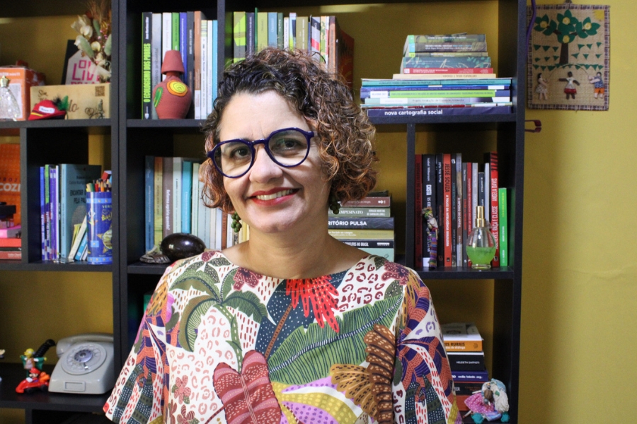 A foto mostra uma mulher de óculos grandes e arredondados, com armação azul-escura, sorrindo para a câmera. Ela tem cabelos curtos e cacheados, com mechas em tom castanho e loiro, e veste uma blusa colorida com estampa vibrante de folhas e formas abstratas. Atrás dela, há uma estante repleta de livros organizados, alguns objetos decorativos e artesanatos