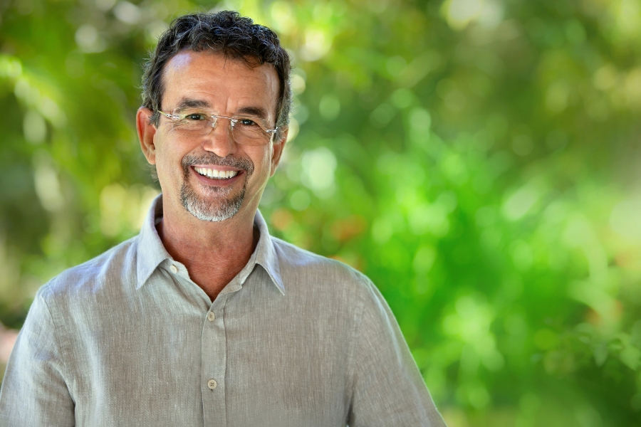 A imagem mostra um homem de pele clara, cabelos curtos e ondulados castanho-escuros com alguns fios grisalhos, barba e bigode bem aparados também grisalhos. Ele usa óculos de armação fina e está sorrindo, transmitindo simpatia. Veste uma camisa de linho bege clara. O fundo está desfocado, mas revela um cenário verde, provavelmente de vegetação iluminada pela luz do dia