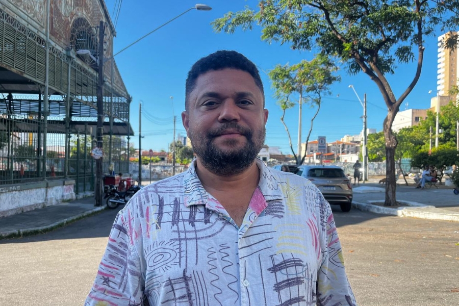 A foto mostra um homem de barba curta e cabelo raspado nas laterais, vestindo uma camisa clara estampada com formas geométricas coloridas. Ele está em pé em uma rua ensolarada, olhando para a câmera com expressão neutra. Ao fundo, há árvores, carros estacionados e construções urbanas