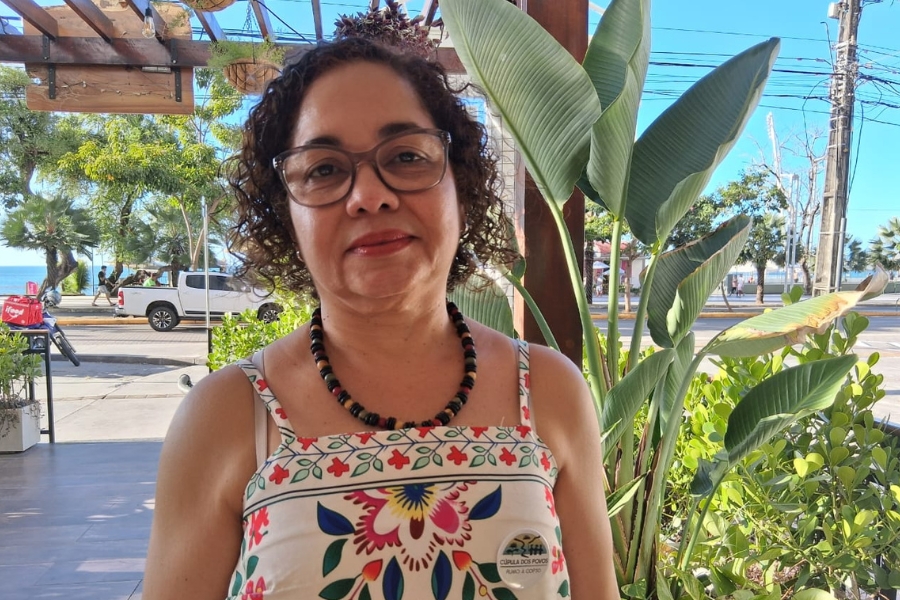 A imagem mostra uma mulher de cabelos escuros e cacheados, usando um colar de sementes e um vestido de alças claro com estampa colorida. Ela está sorrindo levemente, direcionando o olhar para a câmera. Ao, plantas, uma avenida onde passa uma caminhonete branca, árvores e o mar