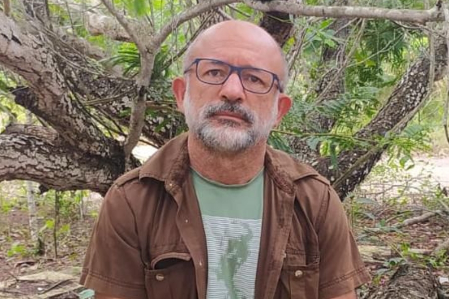 A imagem mostra um homem de pele clara, careca, com barba e bigode grisalhos. Ele usa óculos de armação escura e veste uma camiseta verde com estampa na frente, por baixo de uma camisa marrom de mangas curtas, aberta. O homem está sentado em um ambiente externo, cercado por árvores de troncos retorcidos e folhagem verde, olhando para a câmera com expressão séria