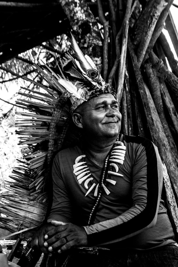A foto em preto e branco mostra um homem indígena sentado, em meio a troncos e galhos grossos. Ele usa um grande cocar de palha e penas, com plumas longas no topo. No pescoço, carrega um colar feito de contas e peças curvas que lembram presas ou ossos. Ele veste uma camisa de mangas compridas e olha para o lado direito da imagem, com as mãos apoiadas sobre as pernas