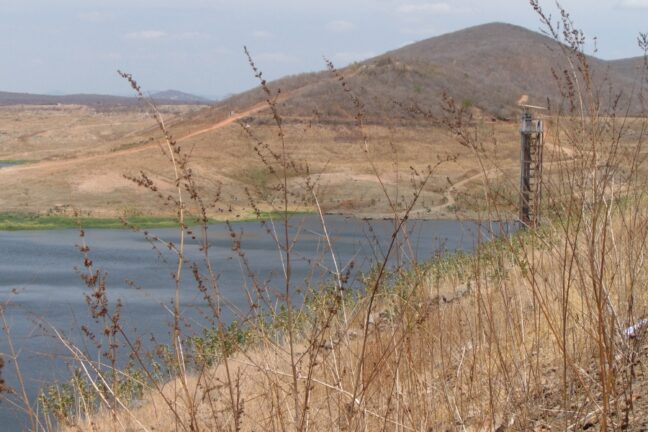 Paisagem de semiárido na seca. Em primeiro plano, galhos secos. Ao fundo, um açude com pouca água, margens áridas e uma torre de concreto. Colinas secas completam a cena sob um céu claro