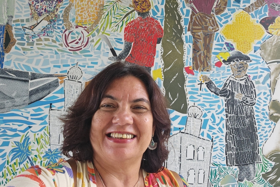 A foto mostra uma mulher de cabelos castanhos escuros e curtos, sorridente, vestindo uma blusa colorida com tons de branco, laranja e amarelo. Ela usa brincos e está em frente a um mural vibrante feito em mosaico, que retrata figuras humanas, elementos da cultura popular, prédios e cores vivas em azul, vermelho, amarelo e verde