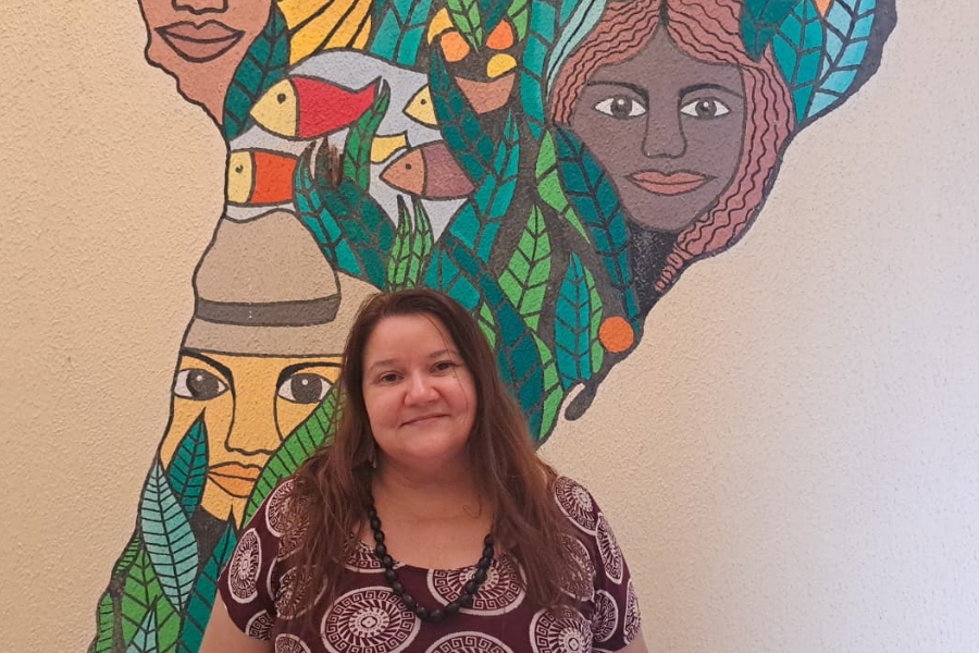 A foto mostra uma mulher de cabelos longos e castanhos, sorrindo levemente para a câmera. Ela veste uma blusa estampada em tons de marrom e branco e um colar preto. Ao fundo, há um mural colorido pintado na parede, representando rostos humanos, peixes e folhas em tons vibrantes de verde, azul, laranja e vermelho, formando uma composição artística que mistura elementos da natureza e da cultura