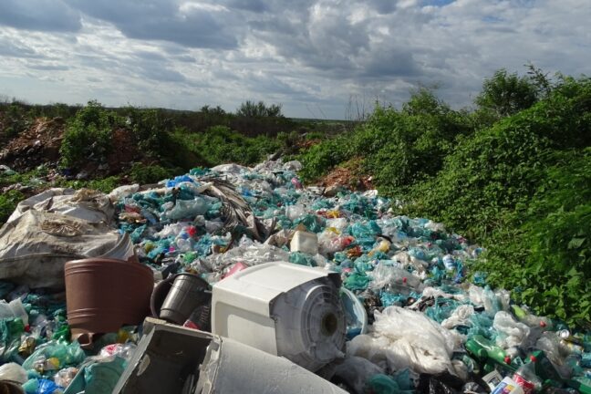 A foto mostra um lixão a céu aberto, onde há grande quantidade de resíduos espalhados pelo chão. Em destaque, vê-se muitas garrafas plásticas amassadas, sacolas e embalagens transparentes e verdes. Também aparecem objetos maiores, como um vaso marrom de plástico e parte de um eletrodoméstico branco descartado. A cena é cercada por vegetação verde densa nas laterais, contrastando com o acúmulo de lixo. No fundo, o céu está parcialmente nublado, com nuvens cinzentas que cobrem boa parte da paisagem