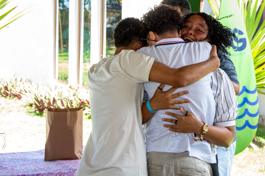 A foto mostra um grupo de quatro pessoas se abraçando de forma muito apertada e emocionada. Uma mulher, de cabelos cacheados e blusa listrada, aparece sorrindo com os olhos marejados, transmitindo alegria. As outras três pessoas estão de costas ou de lado, tornando difícil ver suas expressões. Ao fundo, há um ambiente externo com plantas, uma parede branca com janelas e uma bandeira verde com letras azuis