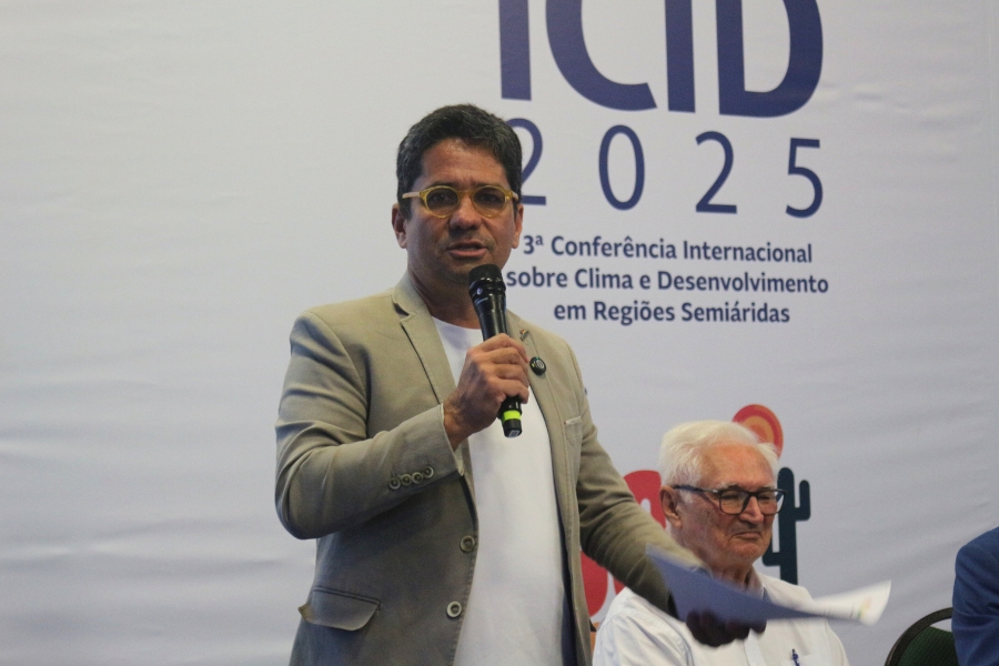 Na foto, um homem está em pé, segurando um microfone e algumas folhas de papel enquanto fala ao público. Ele veste blazer bege claro, camiseta branca e usa óculos de armação amarela. Atrás dele há um painel com a inscrição “ICID 2025 – 3ª Conferência Internacional sobre Clima e Desenvolvimento em Regiões Semiáridas”. Ao fundo, à direita, um homem idoso de cabelos brancos e óculos está sentado, observando a cena