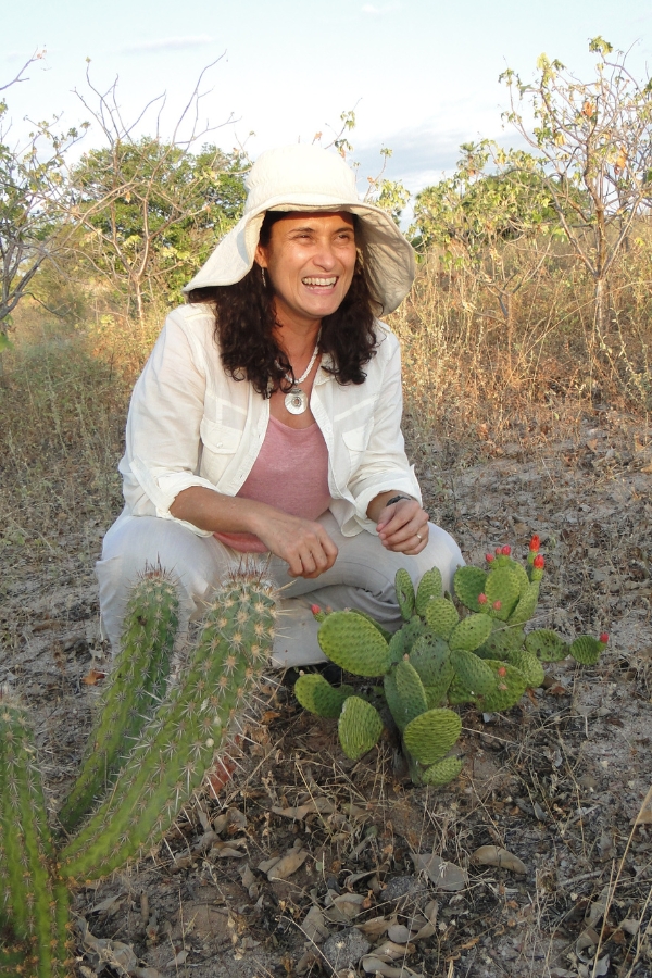 A foto mostra uma mulher sorridente, de cabelos cacheados e longos, vestindo roupas claras e um chapéu grande de aba larga para se proteger do sol. Ela está agachada em meio à vegetação seca da Caatinga, ao lado de dois cactos — um do tipo mandacaru e outro do tipo palma, que tem flores vermelhas na ponta. Ao fundo, aparecem arbustos esparsos típicos do bioma, em um cenário ensolarado e árido