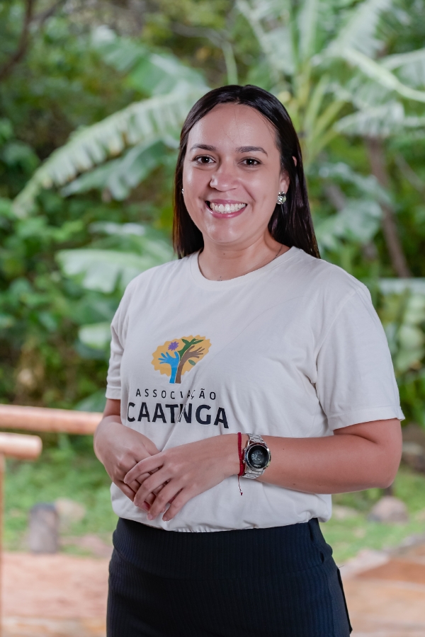 A foto mostra uma mulher de cabelos lisos e longos, soltos, que sorri para a câmera. Ela veste uma camiseta clara com o logotipo da Associação Caatinga estampado no peito e uma calça escura. Usa brincos discretos, um relógio no pulso esquerdo e uma pulseira vermelha no direito. Ao fundo, aparece um cenário verde com folhas largas de bananeiras e vegetação