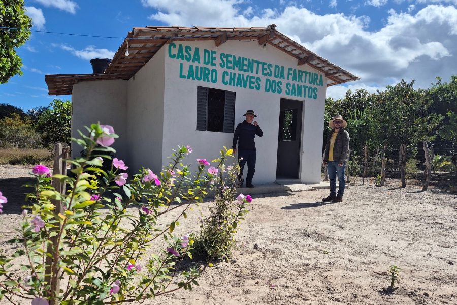A foto mostra uma pequena casa simples de alvenaria pintada de branco, com telhado de telhas de barro, no meio de uma área rural. Acima da porta, em letras verdes, está escrito: “Casa de Sementes da Fartura Lauro Chaves dos Santos”. Em frente ao prédio estão dois homens usando chapéus de palha, um deles vestido de preto e o outro de jaqueta amarela, ambos sorrindo. À esquerda, no primeiro plano, há flores cor-de-rosa. O céu está azul com nuvens brancas, e a casa é cercada por árvores e um cercado rústico de madeira
