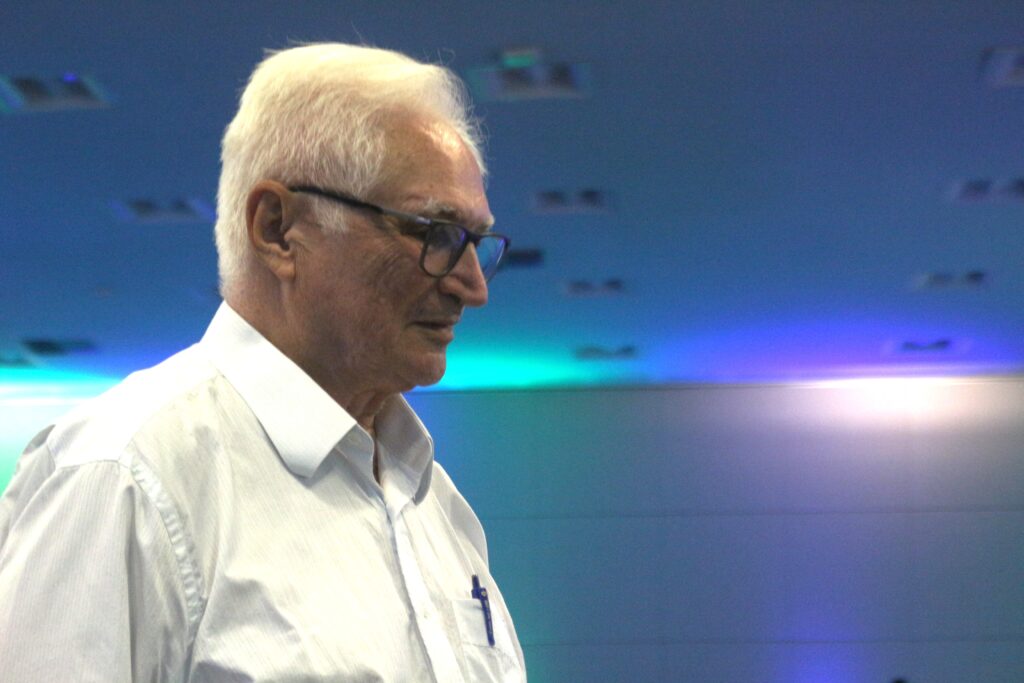 Esta é uma foto em plano médio, tirada em ambiente interno com luz artificial, mostrando o perfil de um homem idoso, de pele clara e cabelos brancos curtos. Ele usa óculos de armação escura e veste uma camisa social branca de manga longa, com um bolso na altura do peito que contém uma caneta de cor escura. O homem está voltado para a direita e sua expressão é séria e concentrada. O fundo é notável pelo uso de iluminação colorida, com tons de azul e roxo projetados em uma parede ou painel liso. O teto, também visível na parte superior, tem um padrão de luminárias embutidas, e a iluminação no ambiente sugere que ele pode estar em uma conferência, evento ou apresentação