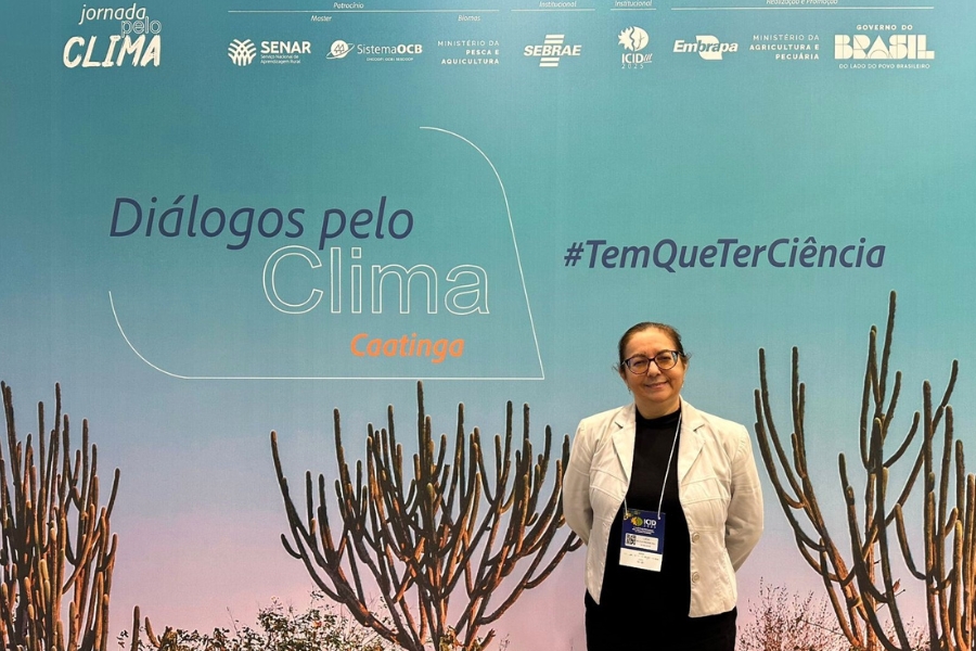 Esta é uma foto de uma mulher de meia-idade, com cabelos escuros presos e usando óculos de grau, que está sorrindo e em pé na frente de um painel de fundo em um evento. Ela veste uma blusa preta, um blazer ou casaco branco e um crachá de identificação pendurado no pescoço. O painel atrás dela, de cor predominantemente azul-claro com transição para tons mais escuros na base, apresenta o título principal "Diálogos pelo Clima" em letras grandes, com a palavra "Caatinga" abaixo, em letras menores, e a hashtag "TemQueTerCiência" à direita. A parte inferior do painel exibe uma ilustração estilizada de galhos de cactos colunares, típicos da Caatinga, que enquadram a mulher. O topo do painel está preenchido com diversos logotipos de patrocinadores e apoiadores, incluindo o Governo do Brasil, SENAR, Sebrae, Embrapa, e diversos ministérios