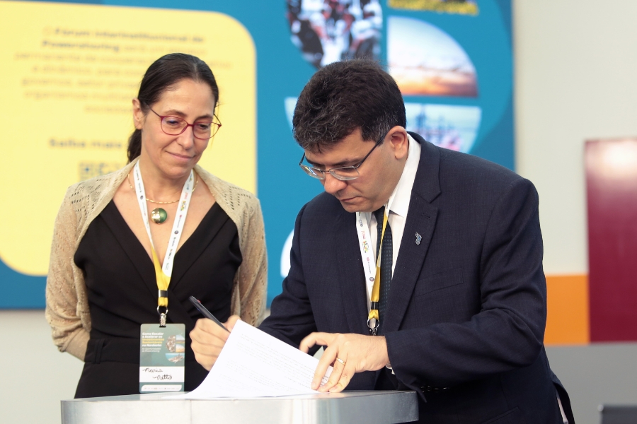 Esta fotografia de close-up, tirada em um evento interno, mostra duas pessoas em um momento de assinatura de documentos. No primeiro plano, à direita, um homem de pele clara e cabelo escuro, vestindo um terno azul-marinho com uma gravata e óculos, está concentrado, curvado enquanto assina um papel em uma superfície clara, segurando uma caneta preta. À esquerda, uma mulher de pele clara, com cabelo escuro preso e usando óculos de aro fino avermelhado, olha para baixo, observando a assinatura. Ela veste um vestido preto e um xale ou casaco de tricô de cor bege claro sobre os ombros, e no pescoço tem um crachá de identificação pendurado, onde se lê o nome "Maria Netto". O fundo é desfocado e colorido em tons de amarelo e azul, com textos e gráficos visíveis