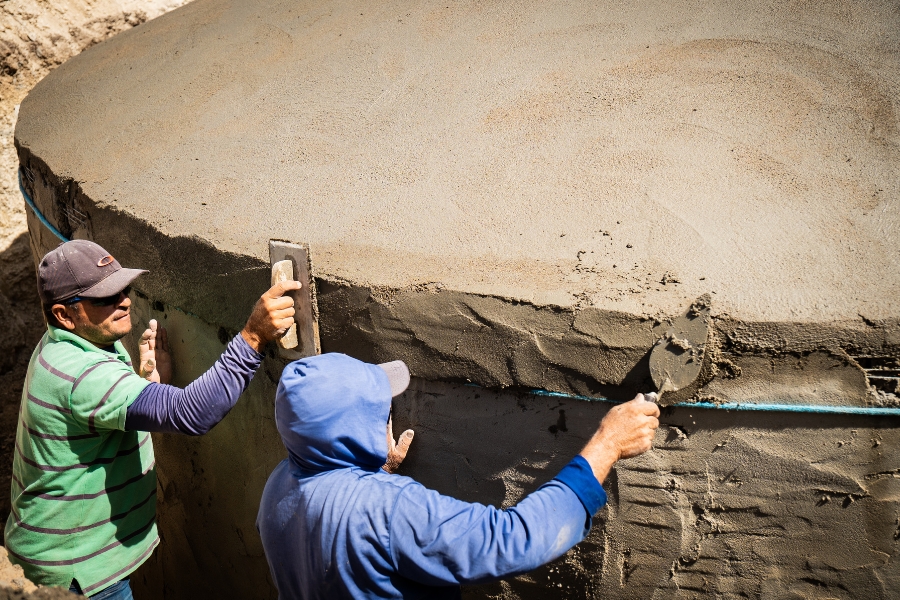 A foto, capturada em plano detalhe, mostra dois homens em processo de construção e reboco de uma grande cisterna de formato arredondado. A superfície da cisterna é de concreto, com a parte inferior e lateral recebendo uma camada de reboco fresco e úmido. O homem à esquerda usa uma camisa polo listrada de verde e cinza, um boné escuro e manguitos longos nos braços; ele aplica o reboco na lateral superior da estrutura com uma desempenadeira retangular. O homem à direita, vestindo um moletom azul com capuz e um boné por baixo, está curvado e aplica o reboco na lateral inferior com uma colher de pedreiro. Um fino cano azul está visível embutido no reboco na lateral da cisterna. O plano de fundo superior revela terra ou argila, sugerindo que a cisterna pode estar sendo construída semienterrada