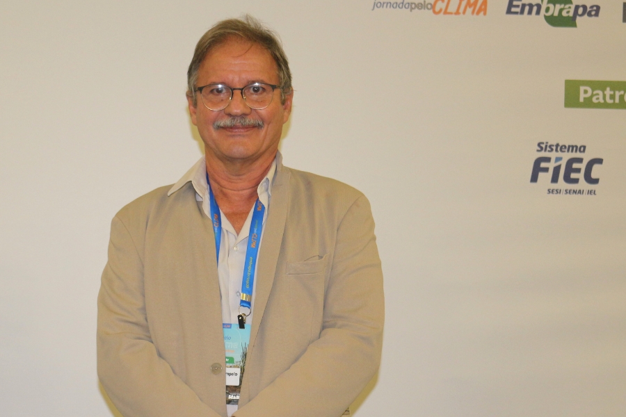 Esta é uma fotografia de meio-corpo, tirada em ambiente interno com boa iluminação, de um homem de meia-idade ou idoso, com cabelos grisalhos e bigode fino. Ele usa óculos de grau de armação clara e está sorrindo discretamente para a câmera. O homem veste uma camisa clara, aberta na gola, e um paletó ou blazer na cor bege ou cáqui. Pendurado em seu pescoço está um crachá de evento preso a uma fita azul. Ele está posicionado em frente a uma parede de fundo branca ou de cor clara, que ocupa a maior parte da composição. No canto superior direito da imagem, há logotipos de patrocinadores, incluindo as palavras "Embrapa" e "Sistema FIEC" com um logotipo relacionado, indicando que a foto foi tirada durante algum evento corporativo ou conferência