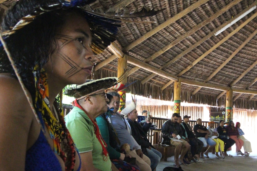Uma fotografia em plano médio, tirada de um ângulo ligeiramente baixo, captura um grupo de pessoas sentadas em uma estrutura aberta, com telhado de palha, que se estende para o lado direito da imagem. Em primeiro plano, no canto esquerdo, uma mulher indígena, com pintura facial tradicional e adornos de penas e contas no cabelo, é vista em perfil, olhando para a direita com uma expressão séria. Ela usa um top azul de crochê ou trançado e seu rosto tem traços de pintura preta geométrica. Atrás dela, uma fila de pessoas sentadas em bancos de madeira se estende horizontalmente. Algumas das pessoas na fila parecem usar cocares tradicionais, e a maioria está vestida com roupas casuais ou sociais. O interior do local é rústico e sombreado, com a luz do dia entrando pelas laterais abertas e iluminando as pessoas. O telhado, feito de palha ou materiais vegetais secos, tem uma estrutura de vigas de madeira visível