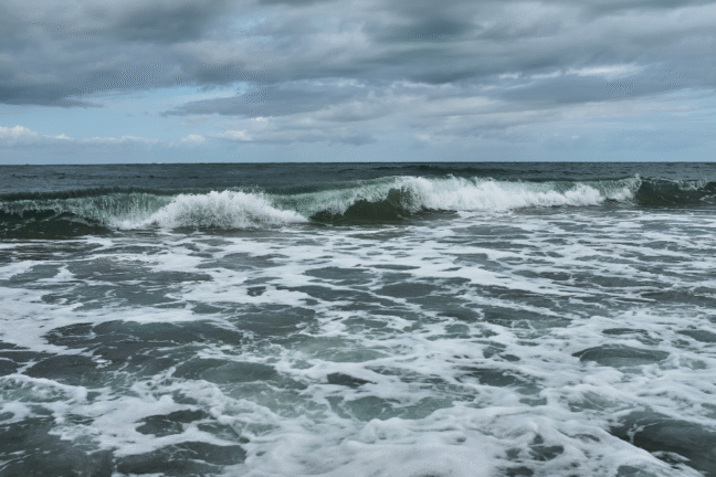 Esta é uma fotografia em close-up de uma onda quebrando no mar sob um céu nublado. A cena é dominada por tons de cinza e azul-marinho. A onda está no meio, espumando em branco enquanto se quebra. A água do mar mais próxima da câmera está agitada e cheia de espuma branca flutuante, cobrindo a parte inferior da imagem. O oceano se estende até o horizonte sob um céu pesado com nuvens escuras e ameaçadoras, indicando um clima tempestuoso ou um dia de pouca luz