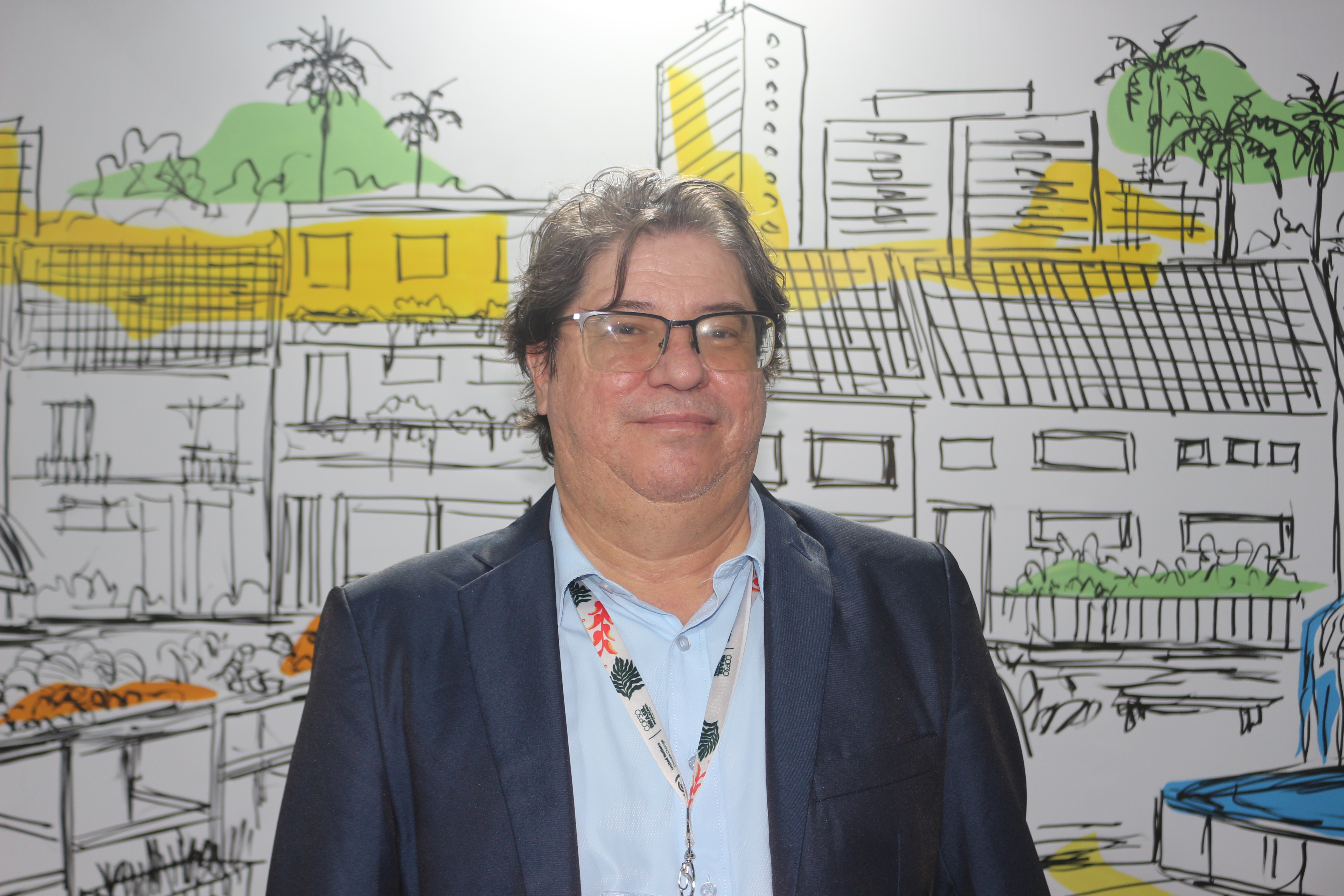 A imagem mostra um homem de meia-idade, de cabelos castanhos e lisos, usando óculos, paletó azul-marinho e camisa azul-clara, com um crachá pendurado no pescoço. Ele está em pé, de frente para a câmera, sorrindo levemente. Ao fundo, há uma ilustração colorida de uma paisagem urbana, com edifícios, árvores e detalhes em amarelo e verde, sugerindo um ambiente moderno e sustentável.