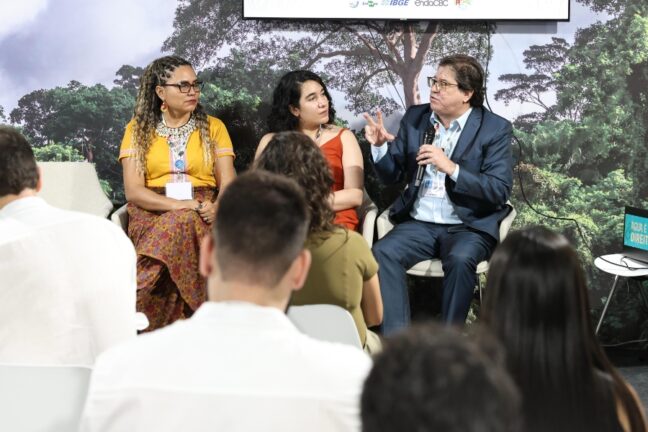 Fotografia mostra três pessoas sentadas em cadeiras no palco de um auditório, participando de um painel de debates diante de uma plateia. À esquerda, uma mulher de tranças longas usa blusa amarela e saia estampada colorida; ao centro, uma mulher de cabelos escuros e curtos veste um vestido laranja; à direita, um homem de terno azul e camisa clara fala ao microfone, gesticulando com a mão. Ao fundo, há uma imagem de floresta tropical e um painel com logotipos institucionais. O público aparece de costas