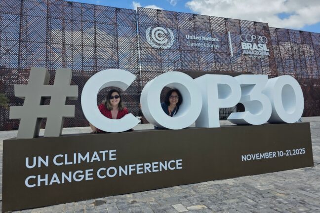 A foto mostra duas mulheres sorrindo atrás de uma grande estrutura tridimensional que forma o texto “#COP30”, instalada ao ar livre sobre um piso de pedras. Elas aparecem enquadradas dentro das letras “O” e “P”, como se estivessem “emolduradas” pelo letreiro. Ao fundo, vê-se a fachada ampla do pavilhão oficial da conferência sobre mudanças climáticas da ONU, com padrões metálicos geométricos e os logotipos da UNFCCC e da COP30