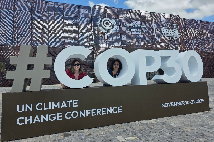A foto mostra duas mulheres sorrindo atrás de uma grande estrutura tridimensional que forma o texto “#COP30”, instalada ao ar livre sobre um piso de pedras. Elas aparecem enquadradas dentro das letras “O” e “P”, como se estivessem “emolduradas” pelo letreiro. Ao fundo, vê-se a fachada ampla do pavilhão oficial da conferência sobre mudanças climáticas da ONU, com padrões metálicos geométricos e os logotipos da UNFCCC e da COP30