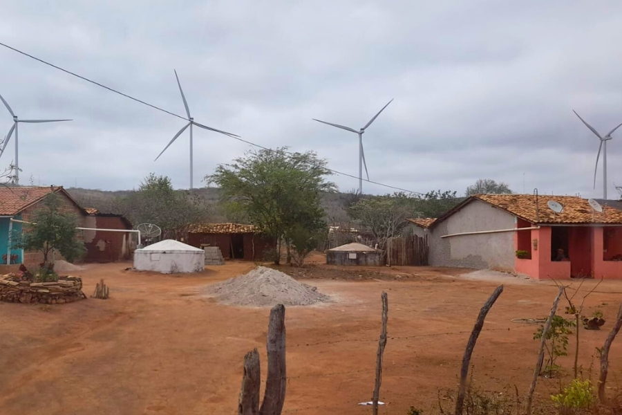 A imagem mostra uma pequena comunidade rural em terreno avermelhado e seco, sob um céu nublado. As casas são simples, feitas de alvenaria e cobertas com telhas de barro. Uma delas é pintada em tom rosa, com uma pessoa sentada à sombra da varanda. No centro do pátio aberto há um monte de areia clara. Cisternas de armazenamento de água, estruturas brancas e cilíndricas, aparecem próximas às moradias. Árvores baixas e arbustos da paisagem semiárida se espalham pelo quintal. Ao fundo, erguidos no horizonte, vários aerogeradores de energia eólica dominam a paisagem. Suas hélices altas contrastam com a escala da comunidade