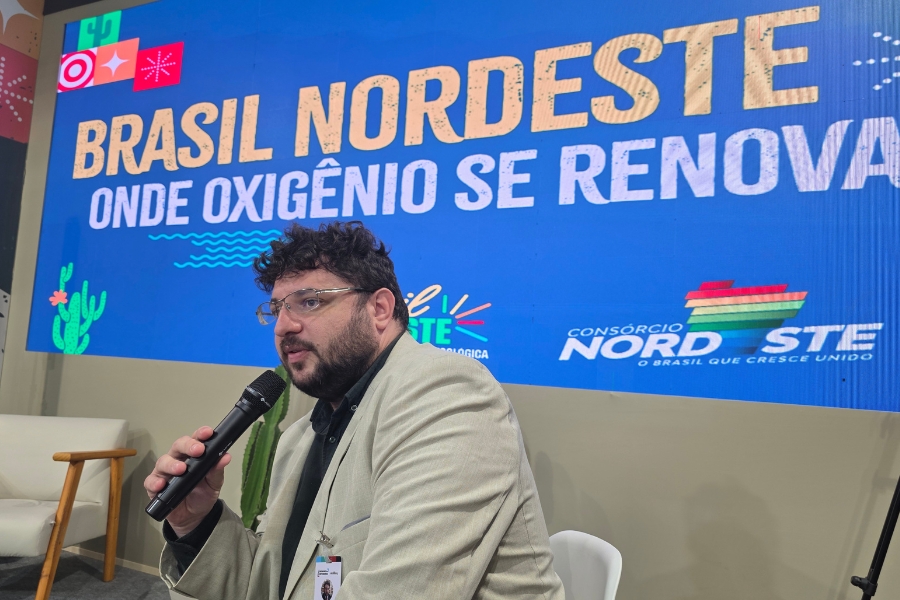 A foto mostra um homem sentado em uma cadeira clara, segurando um microfone enquanto fala. Ele tem barba, cabelos escuros e cacheados, e usa óculos. Veste um blazer bege sobre camisa escura e está posicionado à frente de um grande painel digital azul que traz a frase “Brasil Nordeste: onde oxigênio se renova”, acompanhada de ilustrações de cactos e ondas
