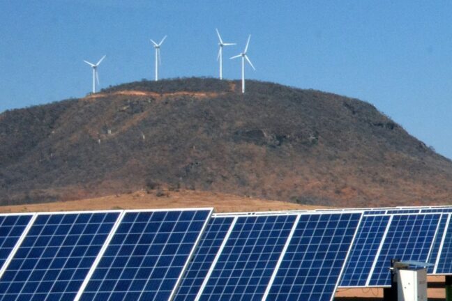 Foto de um cenário de geração de energia renovável em uma paisagem semiárida. No primeiro plano, aparecem fileiras de painéis solares inclinados, ocupando grande parte da base da foto. Ao fundo, sobre uma colina de vegetação seca, cinco aerogeradores eólicos com pás brancas se destacam contra o céu azul.