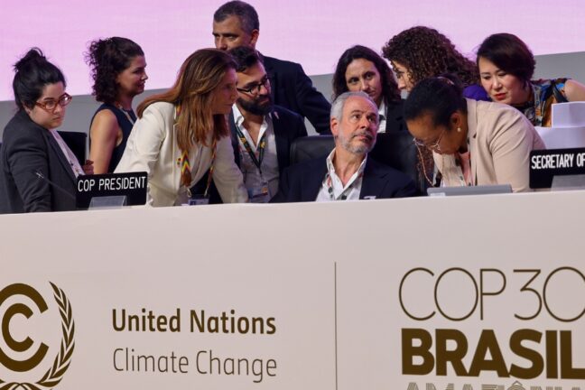 A imagem mostra a mesa principal de uma plenária da COP30, com várias pessoas reunidas em intensa troca de informações antes ou durante uma sessão oficial. No centro, um homem de barba grisalha, sentado, olha atentamente para uma mulher à sua direita, que se inclina para falar com ele, enquanto outra mulher, à esquerda, também se aproxima para conversar. Ao redor deles, outras pessoas — homens e mulheres de diferentes origens — observam, trocam sussurros ou analisam documentos. A mesa exibe placas indicando funções como “COP President” e “Secretary”, e, à gente da mesa, aparece o grande painel oficial da conferência, com os logotipos das Nações Unidas e da COP30 no Brasil