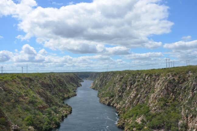 Foto de um cânion profundo por onde corre um largo trecho de rio visto de cima. As paredes rochosas são altas, íngremes e irregulares, cobertas por vegetação típica da Caatinga, em tons de verde seco. O rio, de um azul escuro e calmo, serpenteia pelo centro da cena, contrastando com as encostas pedregosas. No horizonte, o céu ocupa grande parte da imagem, cheio de nuvens brancas e fofas espalhadas por um azul claro luminoso. À esquerda e à direita, no alto das margens, veem-se torres de energia alinhadas, pequenas diante da grandiosidade da paisagem