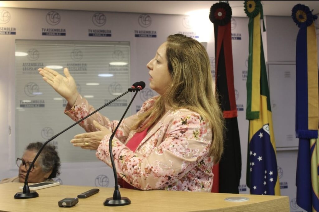 Fotografia mostra uma mulher falando ao microfone em um púlpito, durante um evento em espaço institucional. Ela tem cabelos loiros e longos, veste um blazer claro com estampa floral sobre uma blusa rosa e gesticula com as mãos enquanto fala, em postura firme e expressiva. Atrás dela há bandeiras do Brasil e de outros estados, além de um painel com o logotipo da TV Assembleia Legislativa.