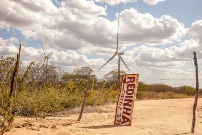 A imagem mostra uma paisagem rural no semiárido, com chão de terra clara, vegetação seca e arbustos espalhados ao longo do terreno. No horizonte, surgem dois aerogeradores brancos, de grandes hélices. No primeiro plano, há uma placa improvisada presa a estacas de madeira, com a inscrição “REDINHA – FORRÓ 27”, pintada em vermelho sobre um fundo claro e já desgastado pelo tempo. Cercas rústicas feitas de arames e troncos delimitam a área.