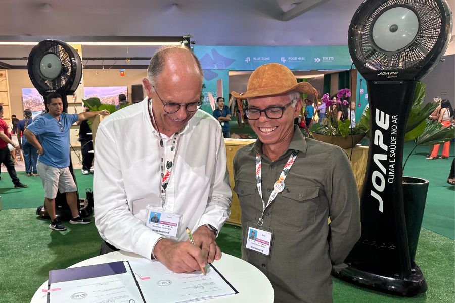 Na foto, dois homens sorriem enquanto participam da assinatura de documentos em uma mesa redonda, dentro de um ambiente movimentado de evento. O homem à esquerda, de camisa branca e óculos, assina o papel com expressão concentrada, enquanto o homem à direita, de camisa verde e chapéu de couro, observa com um sorriso. Ambos usam crachás, indicando participação institucional. Ao fundo, é possível ver pessoas circulando, elementos de decoração e grandes ventiladores industriais, compondo um cenário típico de conferência.