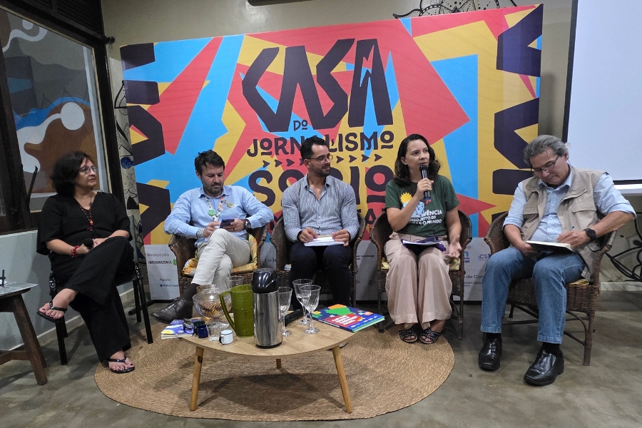 A imagem mostra cinco pessoas sentadas em semicírculo, participando de uma mesa de debate em um ambiente interno. Ao centro do fundo há um painel colorido, em azul, amarelo e vermelho, com letras grandes que formam o nome do evento “Casa do Jornalismo”. À esquerda, uma mulher de óculos, roupas escuras e colar vermelho observa atentamente. Ao lado dela, dois homens, um de camisa clara e outro de listras, folheiam anotações. A quarta pessoa, uma mulher de cabelos castanhos ondulados e camiseta verde, segura um microfone enquanto fala. À direita, um homem de colete claro e jeans lê um caderno apoiado no colo. À frente deles há uma mesa baixa com copos, garrafa térmica e livros